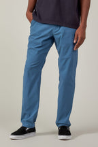 A person wearing the 686 Men's Everywhere® Featherlight Chino Pant - Slim Fit in light blue, paired with a dark t-shirt and black slip-on shoes, stands with one hand in their pocket against a plain background—ideal for summer style.