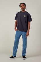 A man stands against a plain background in breathable blue 686 Men's Everywhere® Featherlight Chino Pant - Slim Fit, a loose black T-shirt with a small circular logo, and black slip-on shoes—an effortless summer look with short, braided hair and a relaxed vibe.