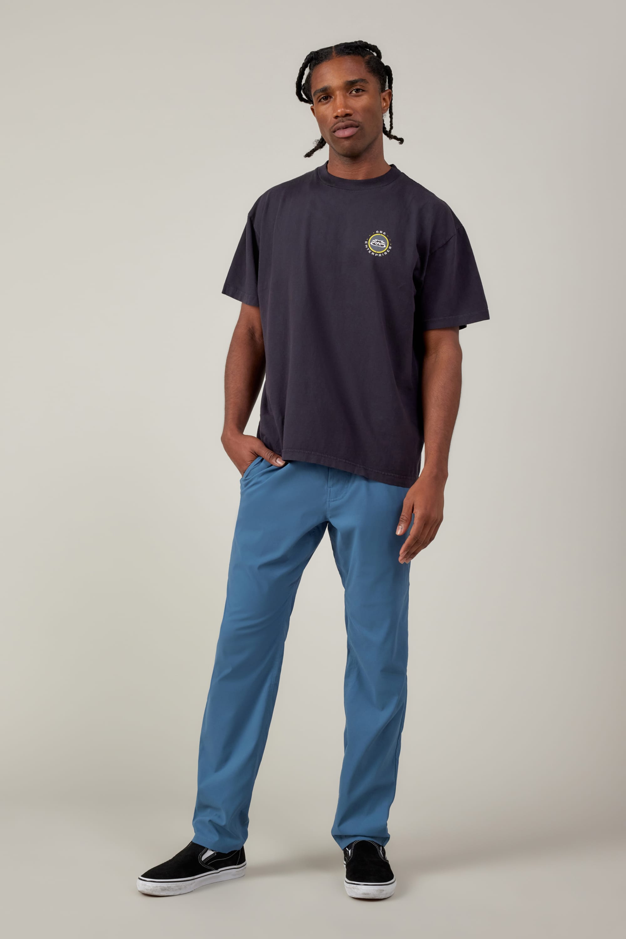 A man stands against a plain background in breathable blue 686 Men's Everywhere® Featherlight Chino Pant - Slim Fit, a loose black T-shirt with a small circular logo, and black slip-on shoes—an effortless summer look with short, braided hair and a relaxed vibe.