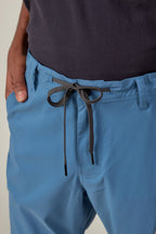 A person wears 686 Men's Everywhere® Featherlight Chino Pant in light blue, featuring a knotted black and white drawstring at the waist for a relaxed, summer-ready look. The slim-fit pants pair with a tucked-in dark shirt.