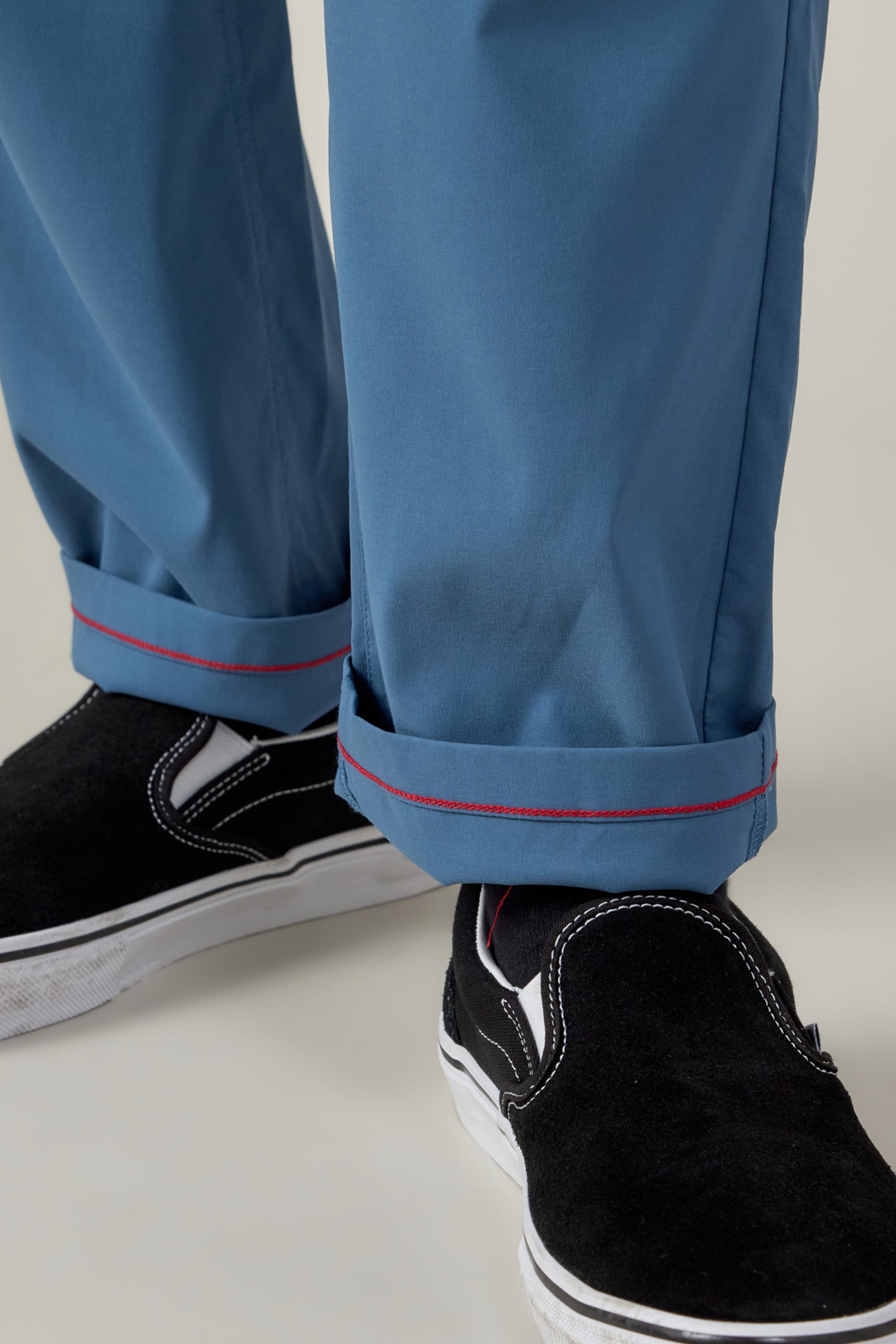 A close-up of a person wearing 686 Men's Everywhere® Featherlight Chino Pant - Slim Fit in light blue with rolled cuffs and red stitching, paired with black slip-on sneakers with white soles—a perfect lightweight choice for summer.