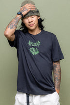 A person with long hair, tattoos, and a beanie stands against a green backdrop wearing the 686 Mens Everywhere drirelease® Tech Tee by 686, featuring a green graphic, paired with light shorts. Their eyes are closed and one hand rests on their forehead.
