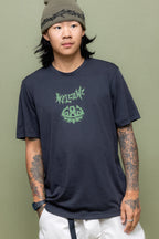 A person with long dark hair and tattoos on both arms wears a gray beanie, light-colored pants, and the 686 Mens Everywhere drirelease® Tech Tee by 686, standing in front of a muted green background.