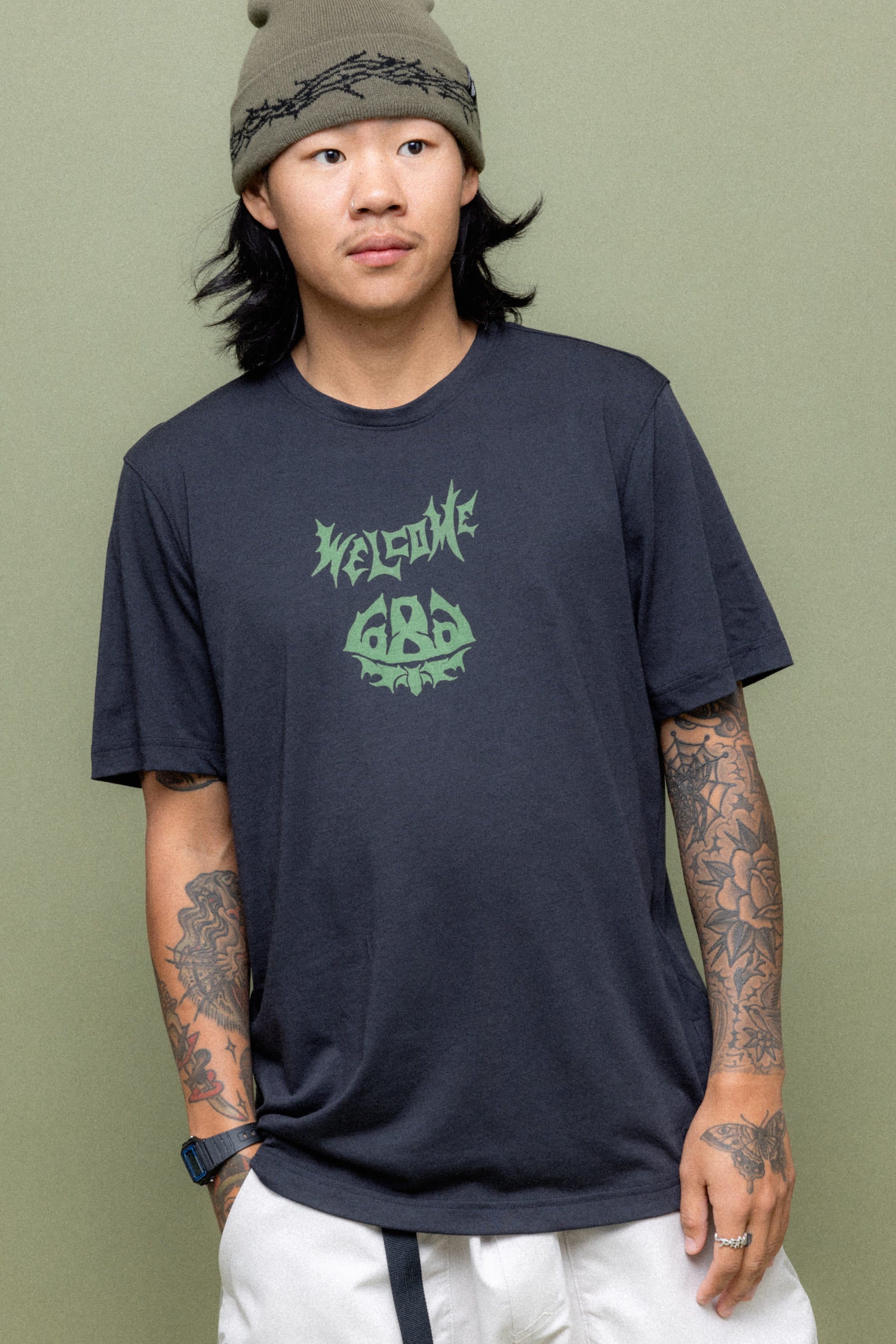 A person with long dark hair and tattoos on both arms wears a gray beanie, light-colored pants, and the 686 Mens Everywhere drirelease® Tech Tee by 686, standing in front of a muted green background.