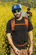 A man in the 686 Mens Everywhere drirelease® Tech Tee stands among yellow wildflowers, wearing a blue cap, sunglasses, and an orange backpack, with another person in dark clothes blurred in the background.