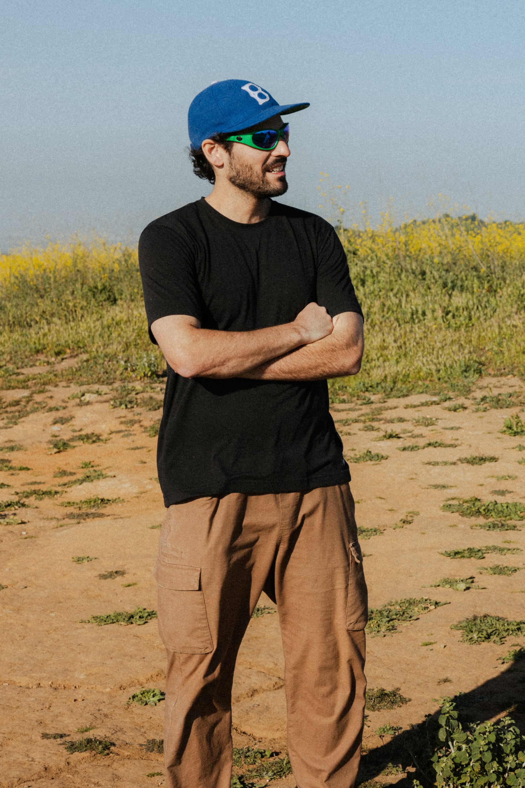 A man wearing the 686 Mens Everywhere drirelease® Tech Tee stands outdoors on a dirt path lined with wildflowers, arms crossed and smiling, with greenery in the background.
