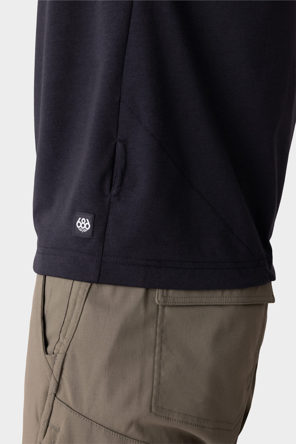 Close-up of a person wearing the 686 Mens Everywhere drirelease® Tech Tee in black, with a small 686 logo tag near the hem. The background is plain and light-colored.