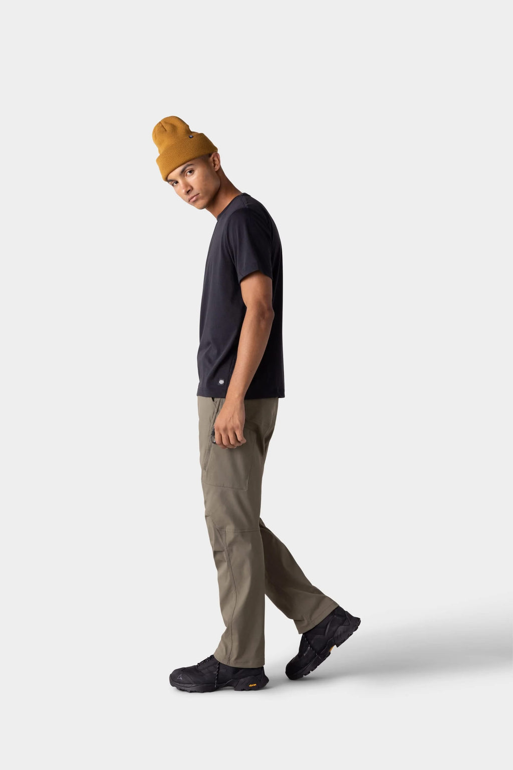 A person in a mustard yellow beanie, olive green pants, black sneakers, and a 686 Mens Everywhere drirelease® Tech Tee by 686 stands against a plain light gray background, looking slightly toward the camera.