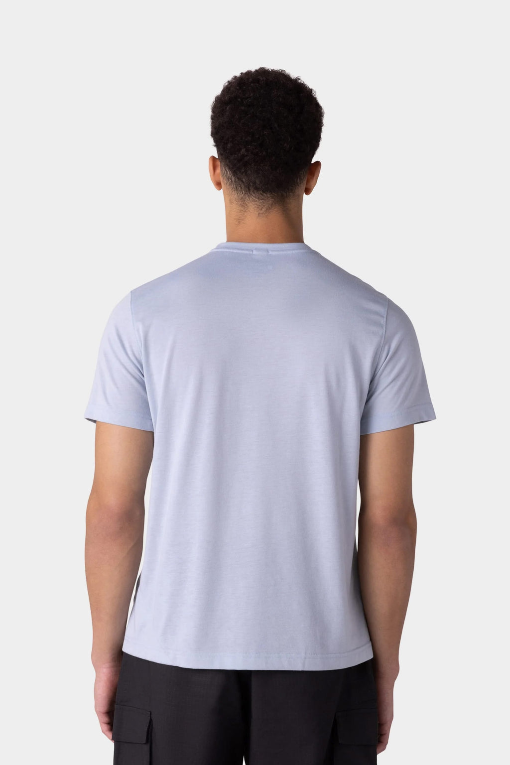 A man with short curly hair is seen from the back, wearing a 686 Mens Everywhere drirelease® Tech Tee in light blue and black pants, standing against a plain light gray background.