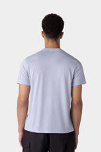 A man with short curly hair is seen from the back, wearing a 686 Mens Everywhere drirelease® Tech Tee in light blue and black pants, standing against a plain light gray background.