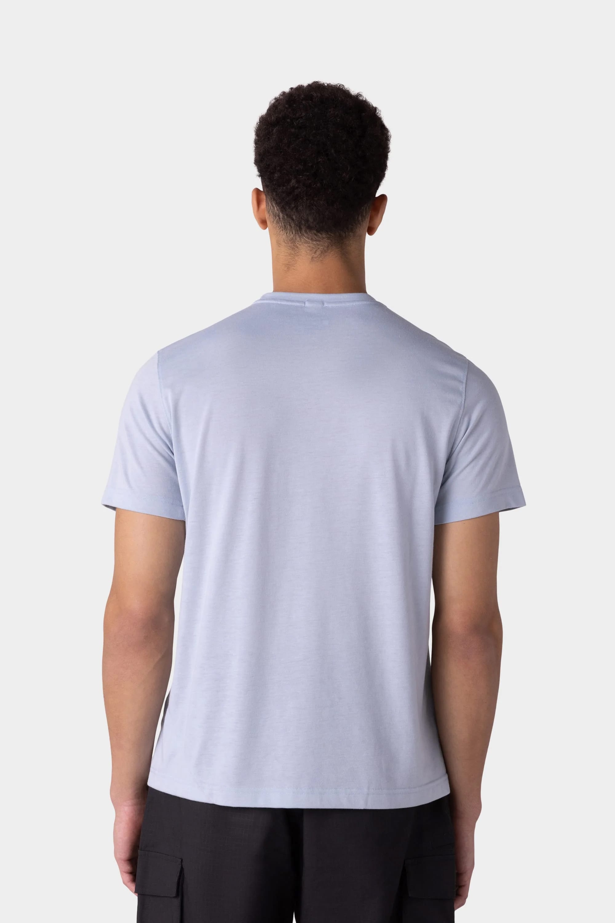 A man with short curly hair is seen from the back, wearing a 686 Mens Everywhere drirelease® Tech Tee in light blue and black pants, standing against a plain light gray background.