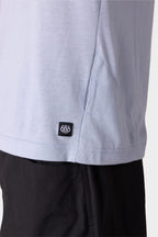 Close-up of the lower side of the 686 Mens Everywhere drirelease® Tech Tee in light gray, featuring a small black and white 686 logo tag near the hem, worn over black pants.