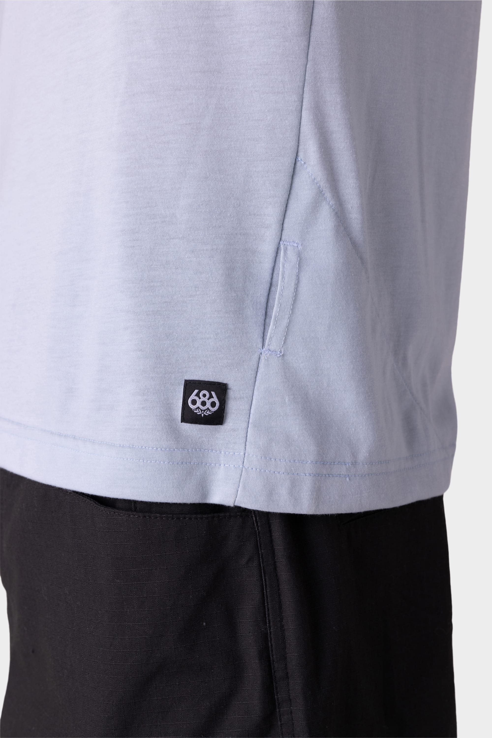 Close-up of the lower side of the 686 Mens Everywhere drirelease® Tech Tee in light gray, featuring a small black and white 686 logo tag near the hem, worn over black pants.