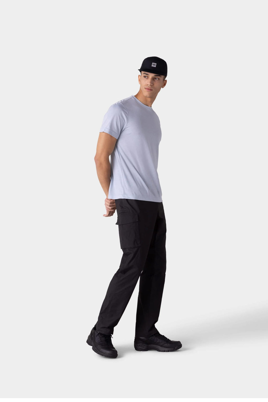 A man in a 686 Mens Everywhere drirelease® Tech Tee, black cargo pants, black sneakers, and a black cap stands against a plain light background, looking to the side with his hands behind his back.