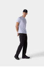 A man in a 686 Mens Everywhere drirelease® Tech Tee, black cargo pants, black sneakers, and a black cap stands against a plain light background, looking to the side with his hands behind his back.