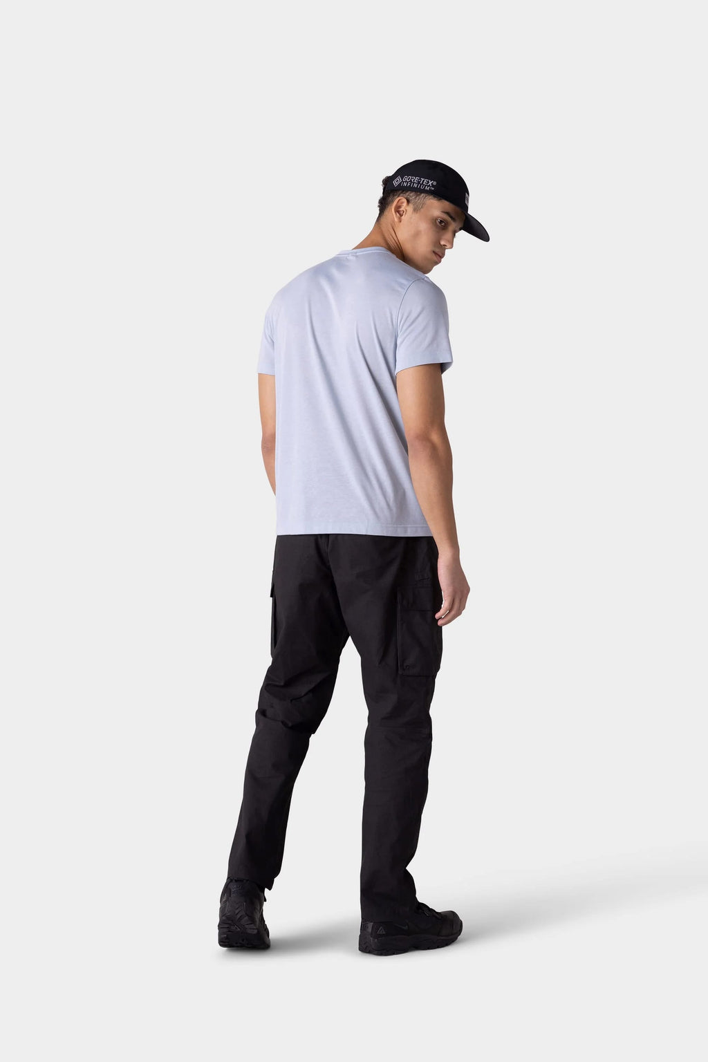 A man faces away, glancing over his shoulder. He wears the 686 Mens Everywhere drirelease® Tech Tee in light gray, with black pants, sneakers, and a cap against a plain light-colored background.