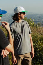 Two people are outdoors on a sunny day. In the foreground, one wears the 686 Mens Everywhere drirelease® Tech Tee by 686, tan pants, a white and green cap, and sunglasses; another in a black shirt and blue cap is partially visible on the left.