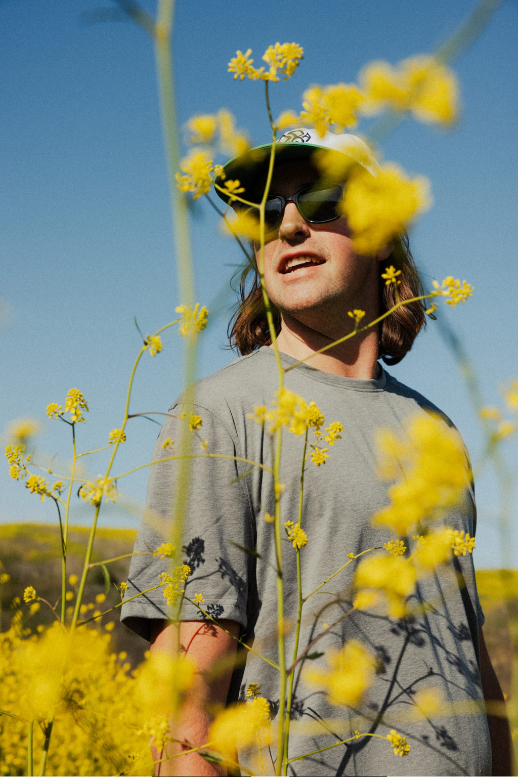 A person wearing the 686 Mens Everywhere drirelease® Tech Tee stands among yellow flowers, smiling under a clear blue sky. Some blooms in the foreground partially obscure the figure. Brand: 686.