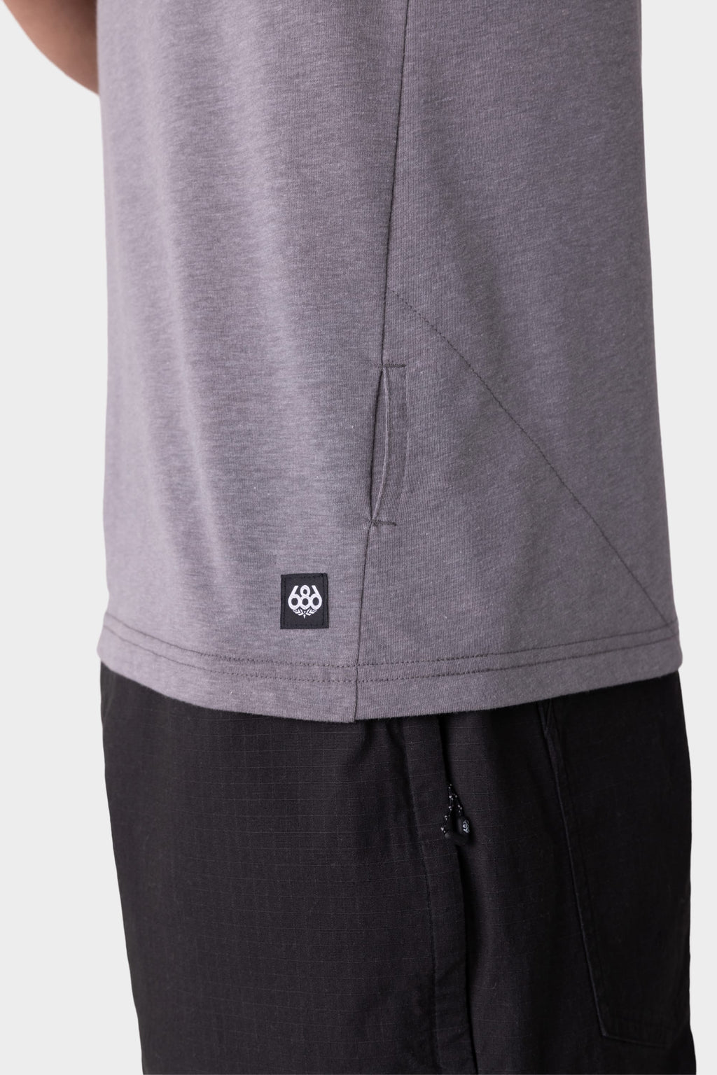 Close-up of a person wearing the 686 Mens Everywhere drirelease® Tech Tee in gray, featuring a small black 686 logo tag near the hem, paired with black pants. Only the lower torso and upper thigh are visible.