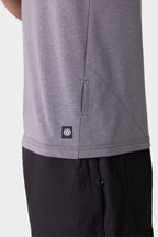 Close-up of a person wearing the 686 Mens Everywhere drirelease® Tech Tee in gray, featuring a small black 686 logo tag near the hem, paired with black pants. Only the lower torso and upper thigh are visible.