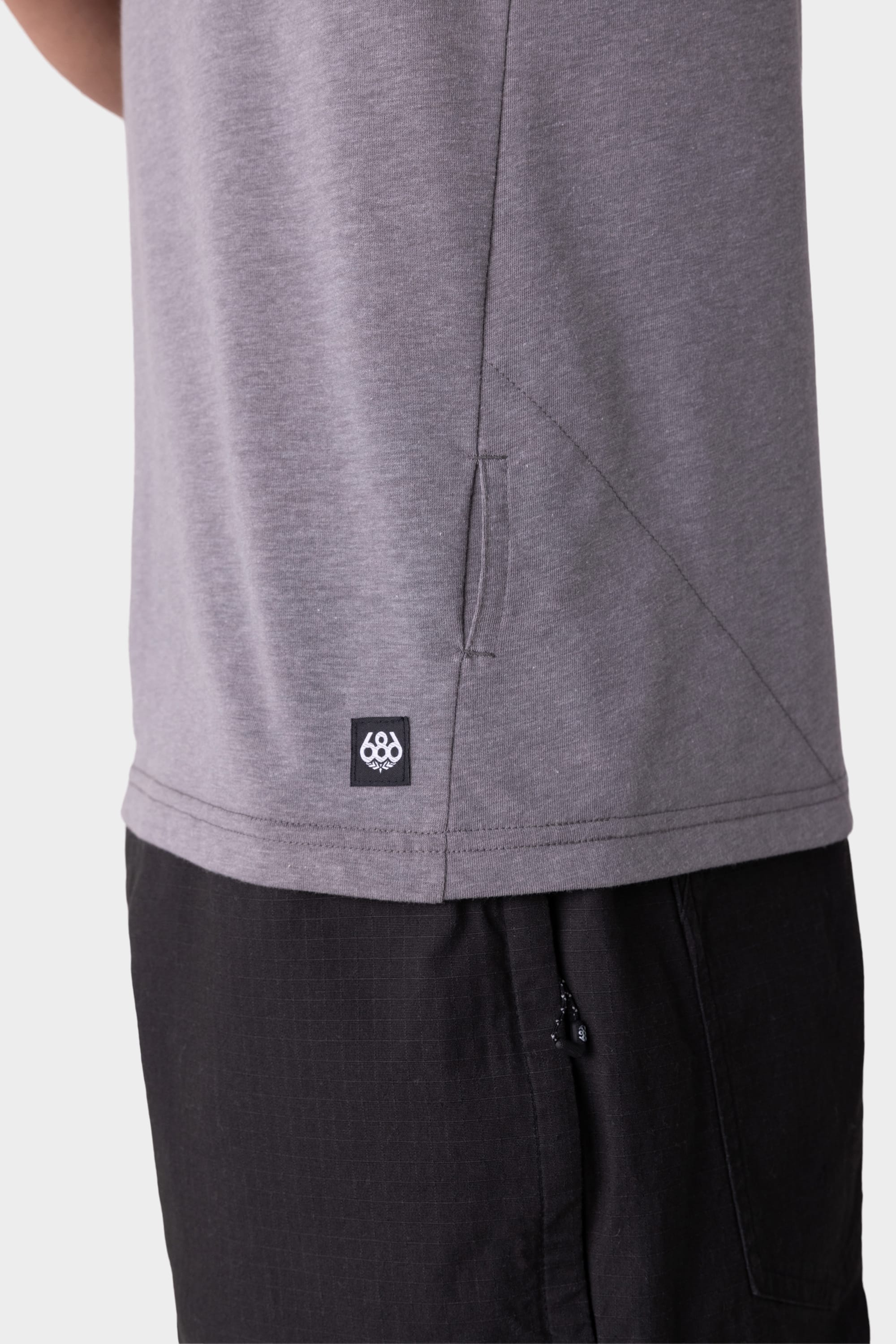 Close-up of a person wearing the 686 Mens Everywhere drirelease® Tech Tee in gray, featuring a small black 686 logo tag near the hem, paired with black pants. Only the lower torso and upper thigh are visible.