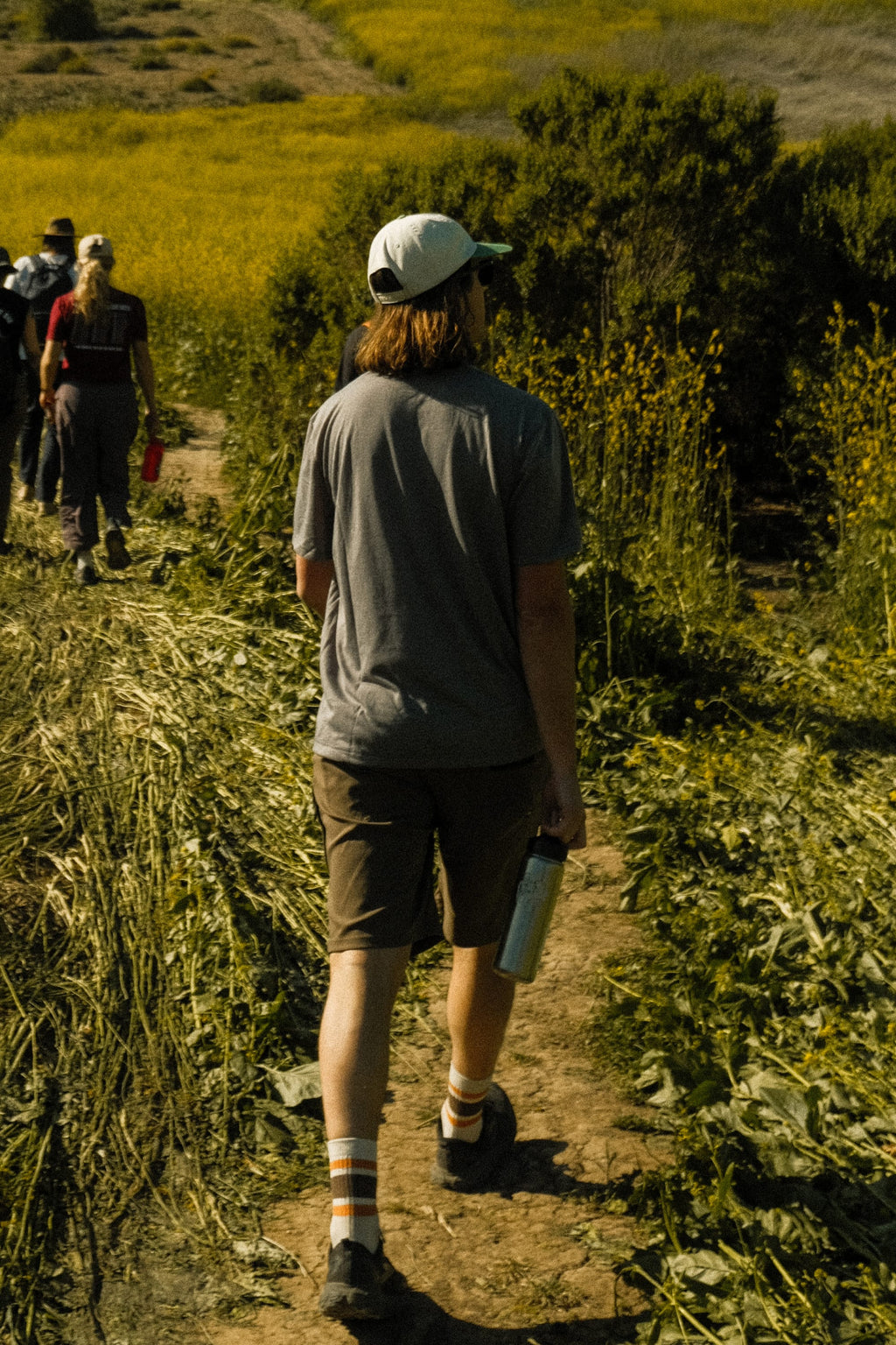 A person wearing the 686 Mens Everywhere drirelease® Tech Tee walks with a group on a dirt trail lined with green plants and yellow wildflowers, holding a water bottle, under the sun.