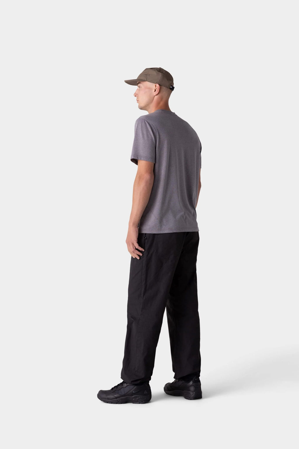 Someone faces away wearing the 686 Mens Everywhere drirelease® Tech Tee in gray, paired with black pants, black sneakers, and a gray cap. The background is plain and light-colored.