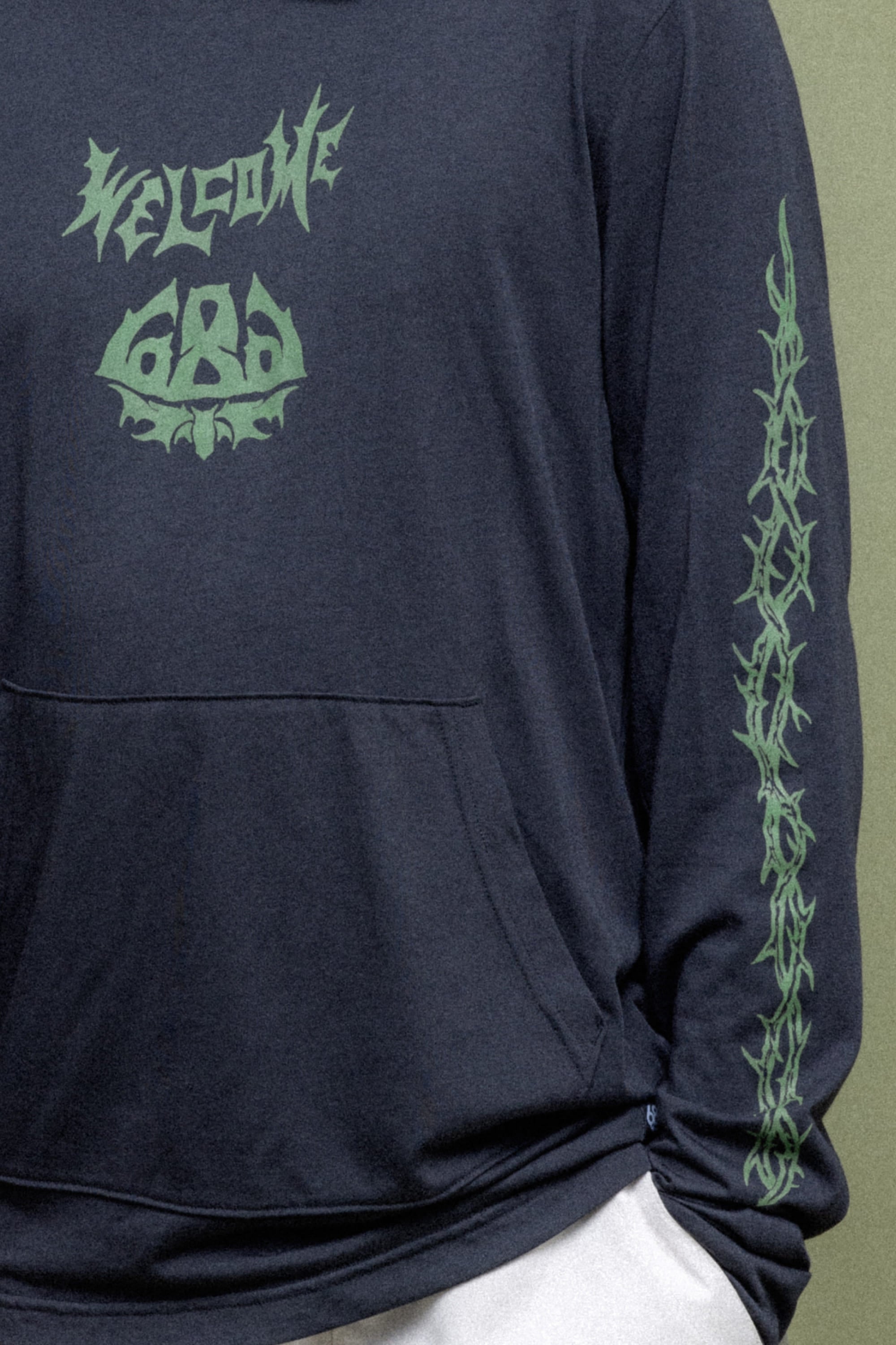 A person wearing the 686 Mens Everywhere drirelease® Long Sleeve Hooded Tech Tee with green Welcome 666 text on the chest and matching sleeve graphics, hand in pocket. Brand: 686.