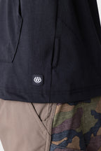 Close-up of a person wearing the 686 Mens Everywhere drirelease® Long Sleeve Hooded Tech Tee in black, featuring a small 686 logo tag near the hem, paired with pants that have solid tan and camouflage panels.