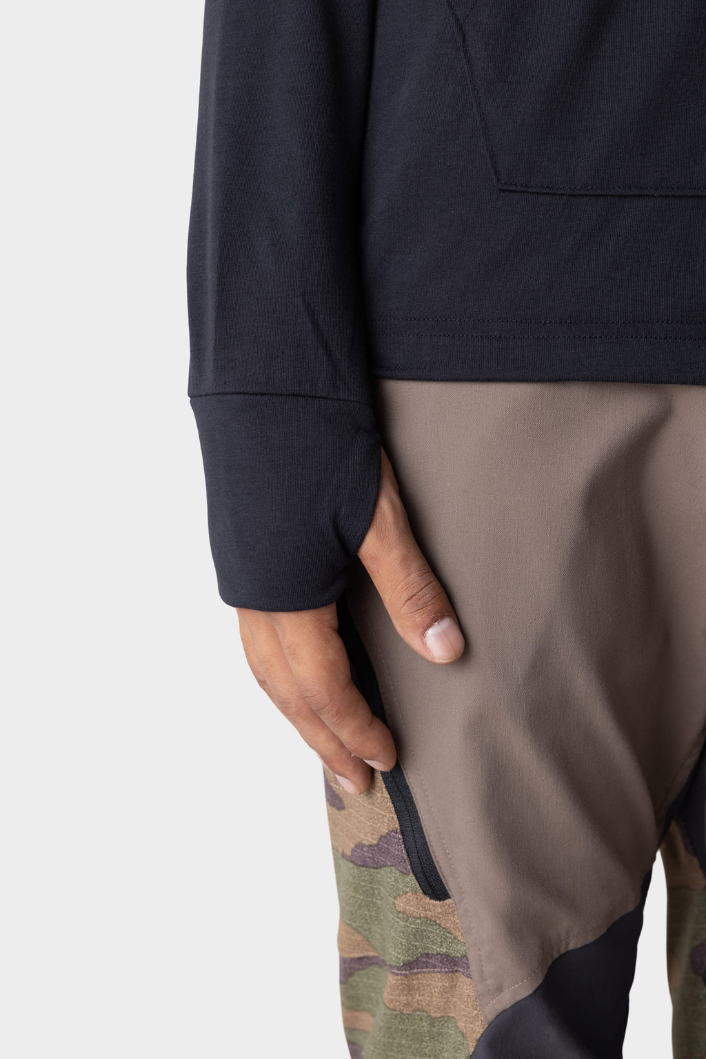 A close-up shows a person’s left hand by their side, wearing the 686 Mens Everywhere drirelease® Long Sleeve Hooded Tech Tee in dark color, paired with pants featuring brown and camouflage panels against a plain light gray background.