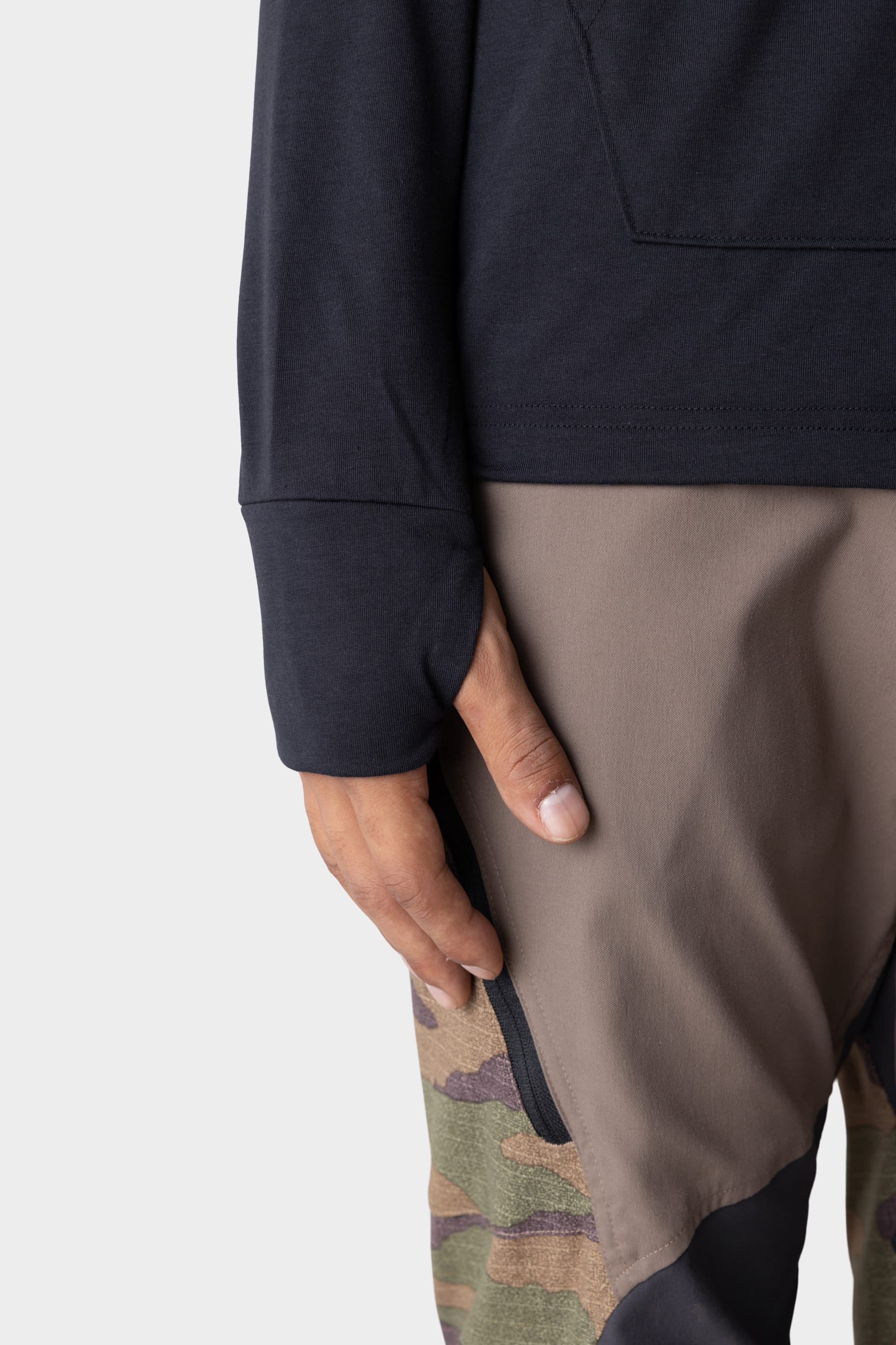 A close-up shows a person’s left hand by their side, wearing the 686 Mens Everywhere drirelease® Long Sleeve Hooded Tech Tee in dark color, paired with pants featuring brown and camouflage panels against a plain light gray background.