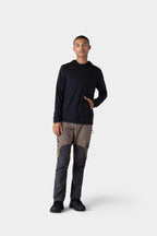 A person stands against a light gray background, wearing the 686 Mens Everywhere drirelease® Long Sleeve Hooded Tech Tee in black, with brown and gray color-blocked pants and black shoes, one hand in the hoodie’s front pocket.
