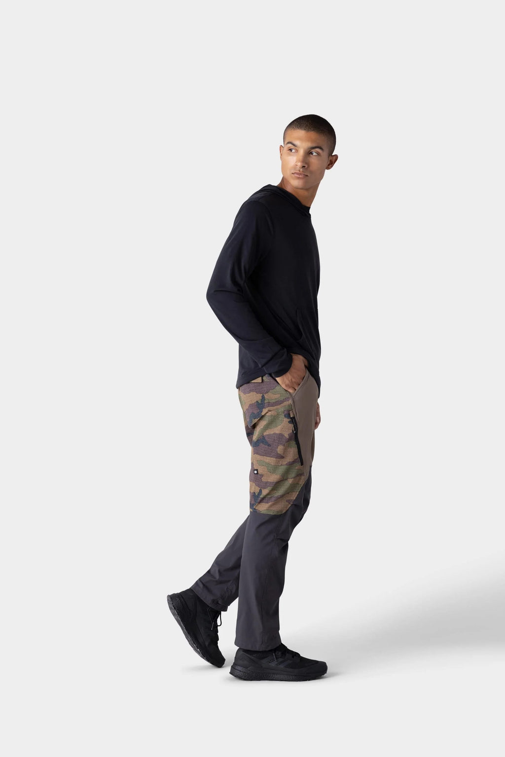 A person stands against a plain light gray background, looking to the side. They wear the 686 Mens Everywhere drirelease® Long Sleeve Hooded Tech Tee by 686, camouflage cargo pants with dark gray panels, and black shoes.