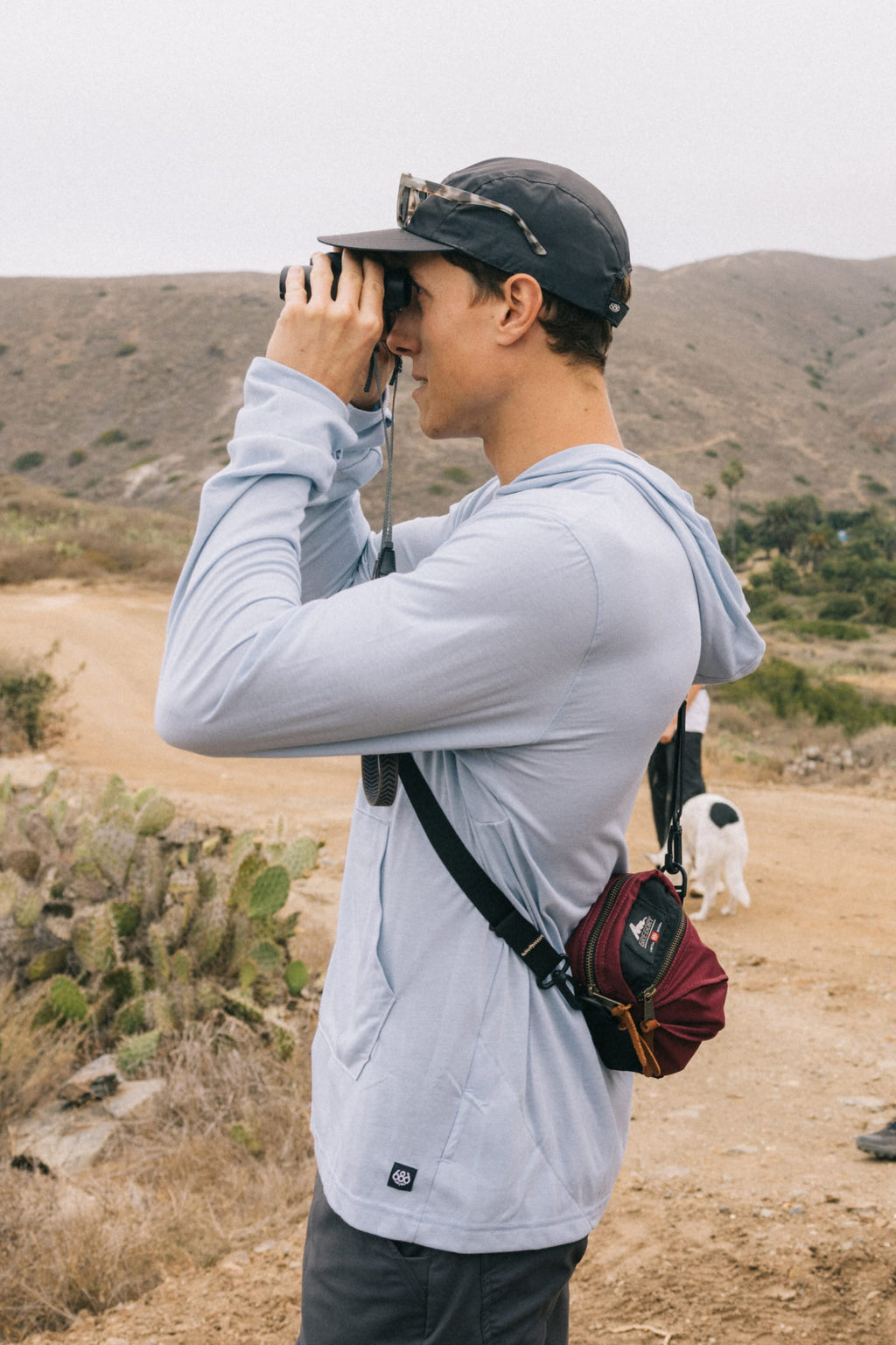 A person wears the 686 Mens Everywhere drirelease® Long Sleeve Hooded Tech Tee in light blue, along with a cap and sunglasses, while looking through binoculars on a desert trail. A small black and white dog is seen in the background.
