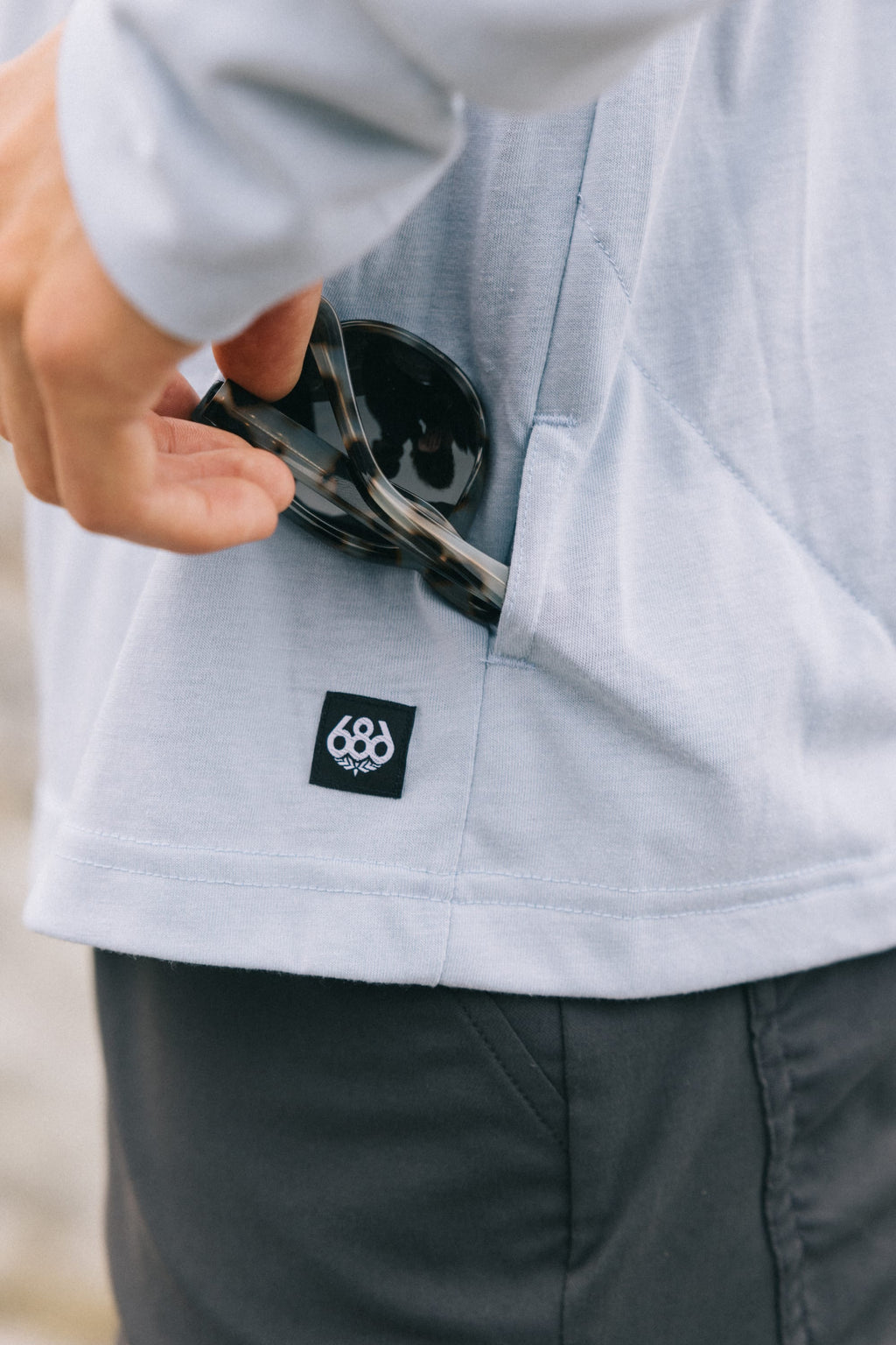 A person in the 686 Mens Everywhere drirelease® Long Sleeve Hooded Tech Tee (light gray) puts tortoiseshell sunglasses into a side pocket. The shirt’s side pocket features a small black and white 686 logo tag.