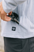 A person in the 686 Mens Everywhere drirelease® Long Sleeve Hooded Tech Tee (light gray) puts tortoiseshell sunglasses into a side pocket. The shirt’s side pocket features a small black and white 686 logo tag.