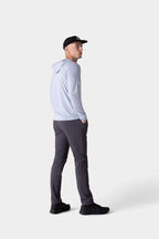 A man wears the 686 Mens Everywhere drirelease® Long Sleeve Hooded Tech Tee by 686 with dark gray pants, black shoes, and a black cap, standing with hands in pockets against a plain light background.