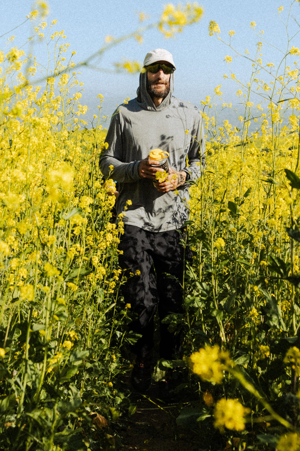 Wearing the 686 Mens Everywhere drirelease® Long Sleeve Hooded Tech Tee, a person in sunglasses and a white cap walks through tall yellow wildflowers with a yellow can under clear, sunny skies.