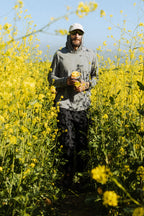 Wearing the 686 Mens Everywhere drirelease® Long Sleeve Hooded Tech Tee, a person in sunglasses and a white cap walks through tall yellow wildflowers with a yellow can under clear, sunny skies.
