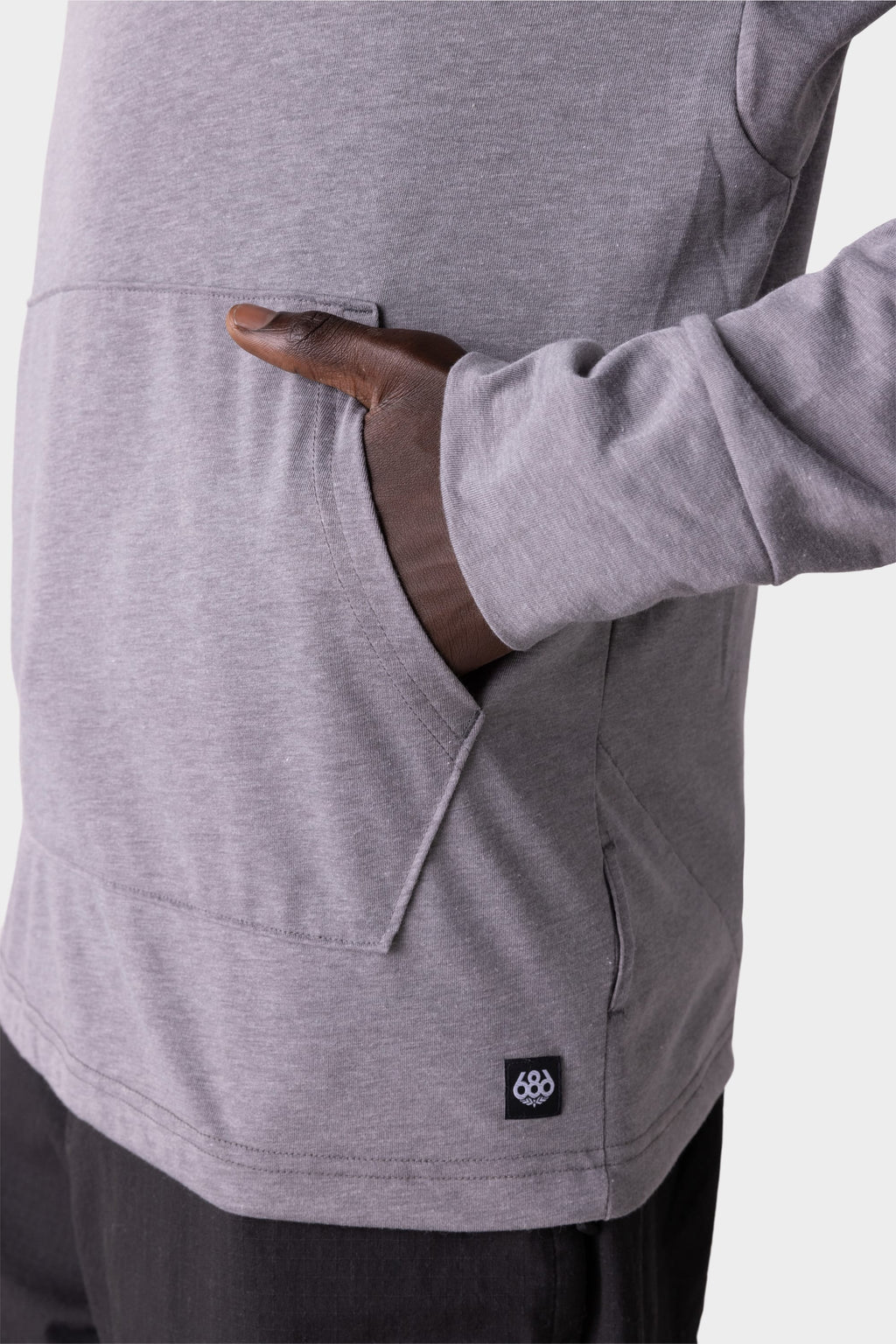A person wears the 686 Mens Everywhere drirelease® Long Sleeve Hooded Tech Tee in light gray, right hand in the front pocket. A small black tag with a white logo is near the pocket, set against a plain, light-colored background.