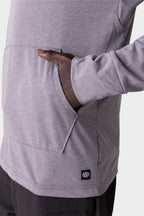 A person wears the 686 Mens Everywhere drirelease® Long Sleeve Hooded Tech Tee in light gray, right hand in the front pocket. A small black tag with a white logo is near the pocket, set against a plain, light-colored background.
