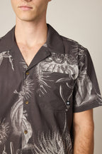 A man wears the 686 Men's Nomad Perforated Button Down Shirt—a dark, lightweight shirt with a white botanical and cactus print, chest pocket, and brown buttons. Only his upper torso and part of his face are visible.