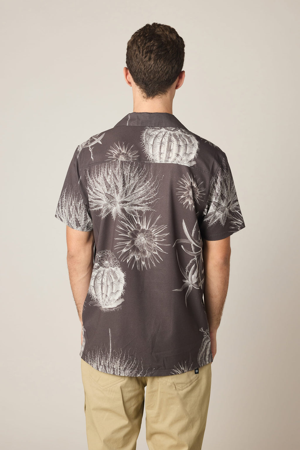 A man, seen from behind against a plain background, wears the 686 Men's Nomad Perforated Button Down Shirt by 686—a dark lightweight shirt featuring a white cactus print—paired with light-colored pants.