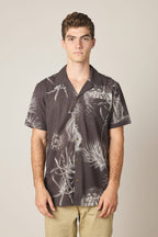 A young man with short curly hair wears the 686 Men's Nomad Perforated Button Down Shirt by 686, featuring a dark, lightweight design with a white floral pattern and beige pants, standing against a plain light background.