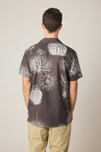 A man faces away wearing the 686 Men's Nomad Perforated Button Down Shirt by 686, a lightweight short-sleeve shirt with a white cactus print, paired with beige pants against a plain light background.