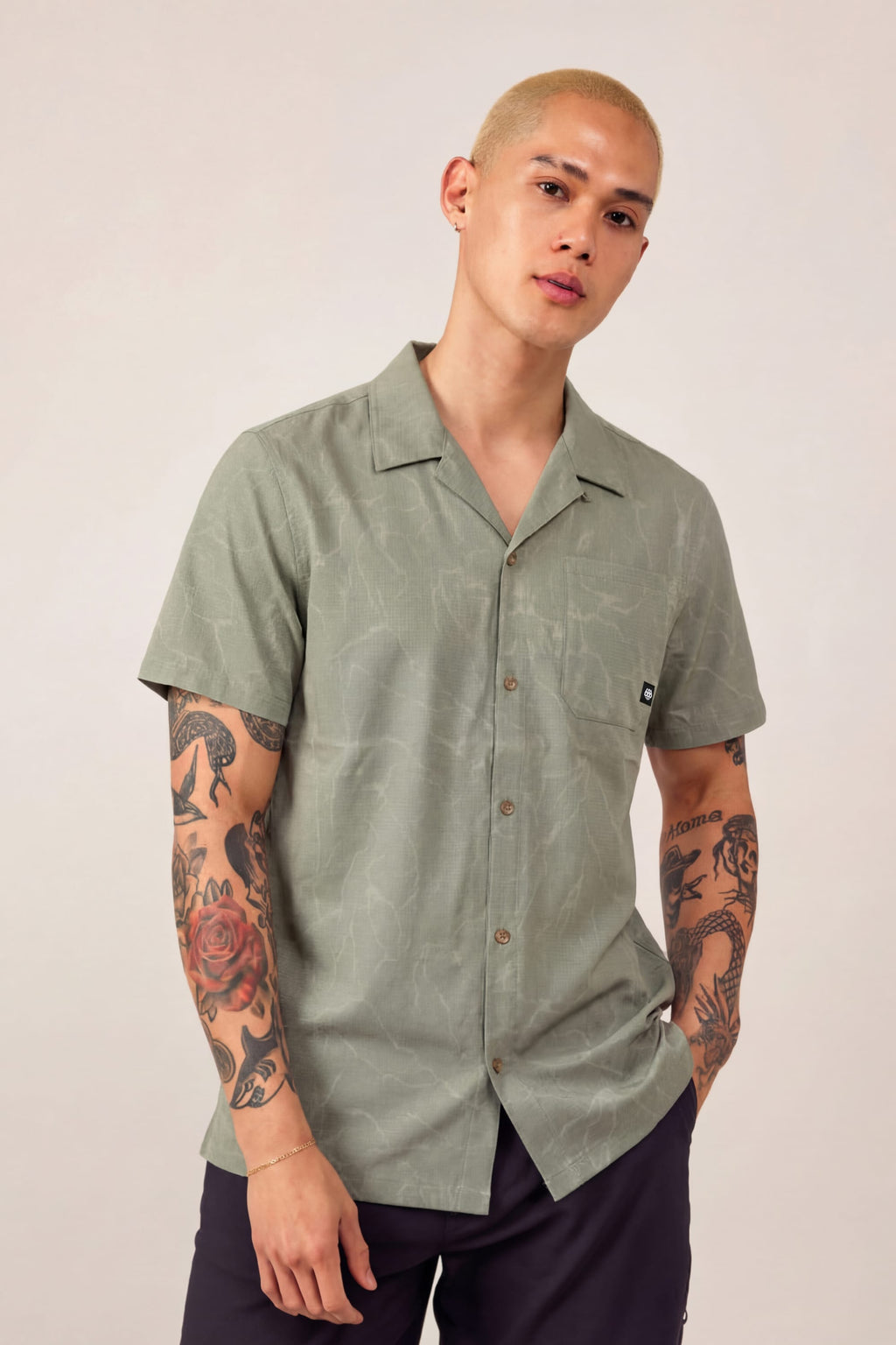 A person with short blond hair and visible arm tattoos wears the 686 Men's Nomad Perforated Button Down Shirt in light green and dark pants, standing with one hand in their pocket against a plain background.