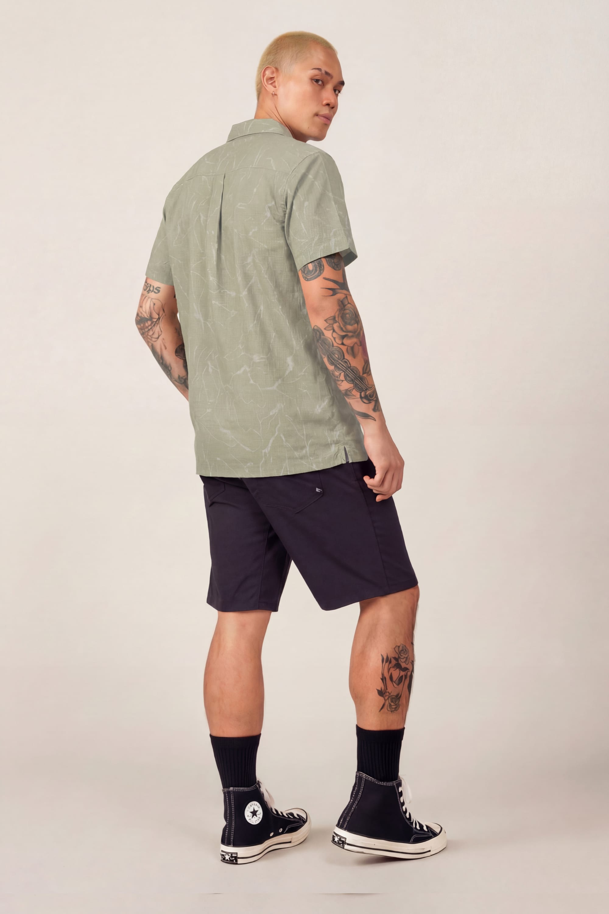 A person with short blond hair and visible tattoos stands facing away, looking over their shoulder. They wear the 686 Men's Nomad Perforated Button Down Shirt in sage green, black shorts, black socks, and black Converse sneakers.