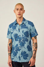 A person with short blond hair and arm tattoos wears the 686 Men's Nomad Perforated Button Down Shirt by 686, featuring a bold dark blue floral pattern, standing facing forward against a plain, light-colored background.