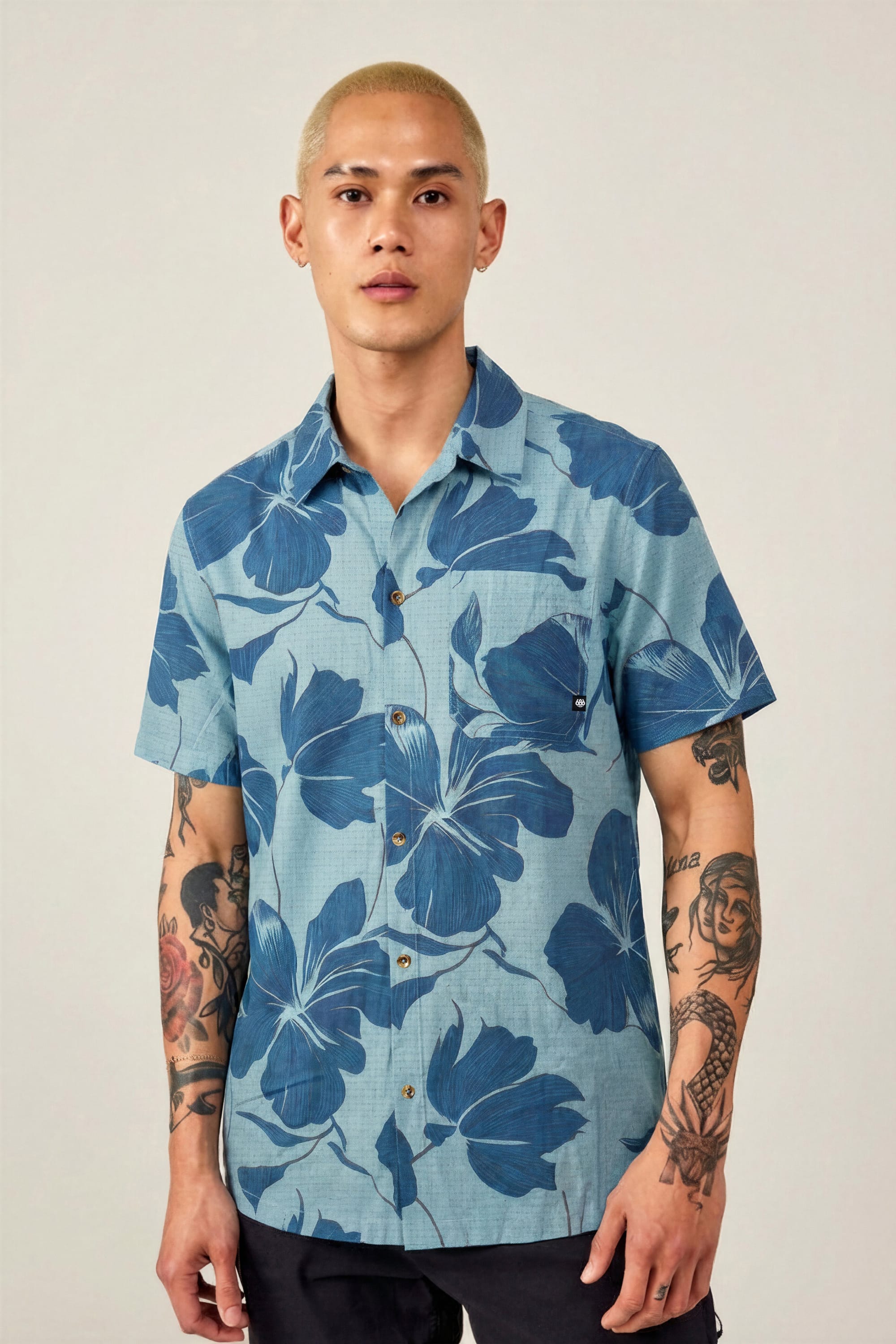 A person with short blond hair and tattooed arms wears the 686 Men's Nomad Perforated Button Down Shirt by 686, featuring a blue lightweight fabric with a bold dark blue floral pattern, against a plain light background.