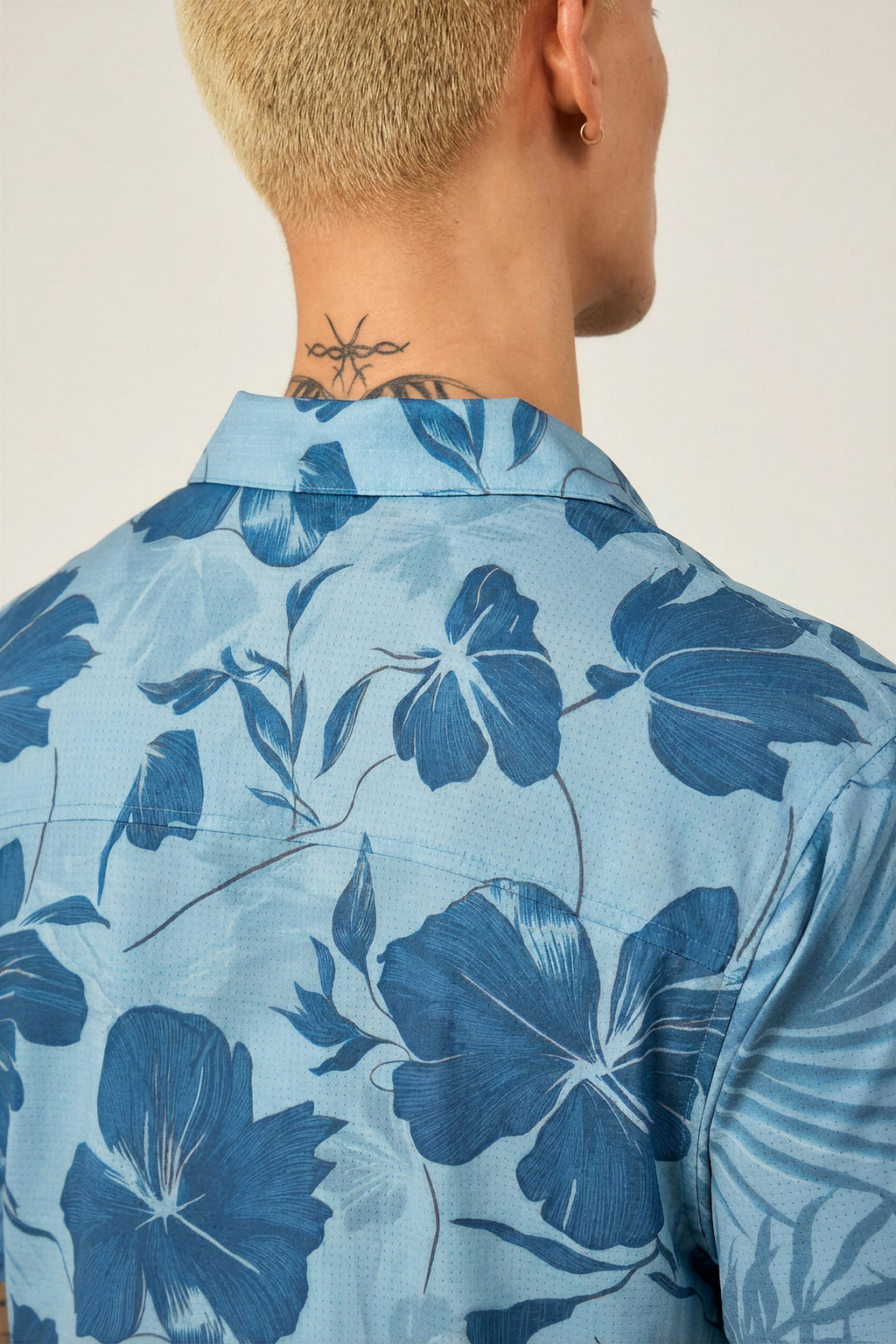 A person with short, light blond hair and a neck tattoo is seen from behind wearing the 686 Men's Nomad Perforated Button Down Shirt by 686, featuring a lightweight, moisture-wicking fabric and a dark blue floral pattern.